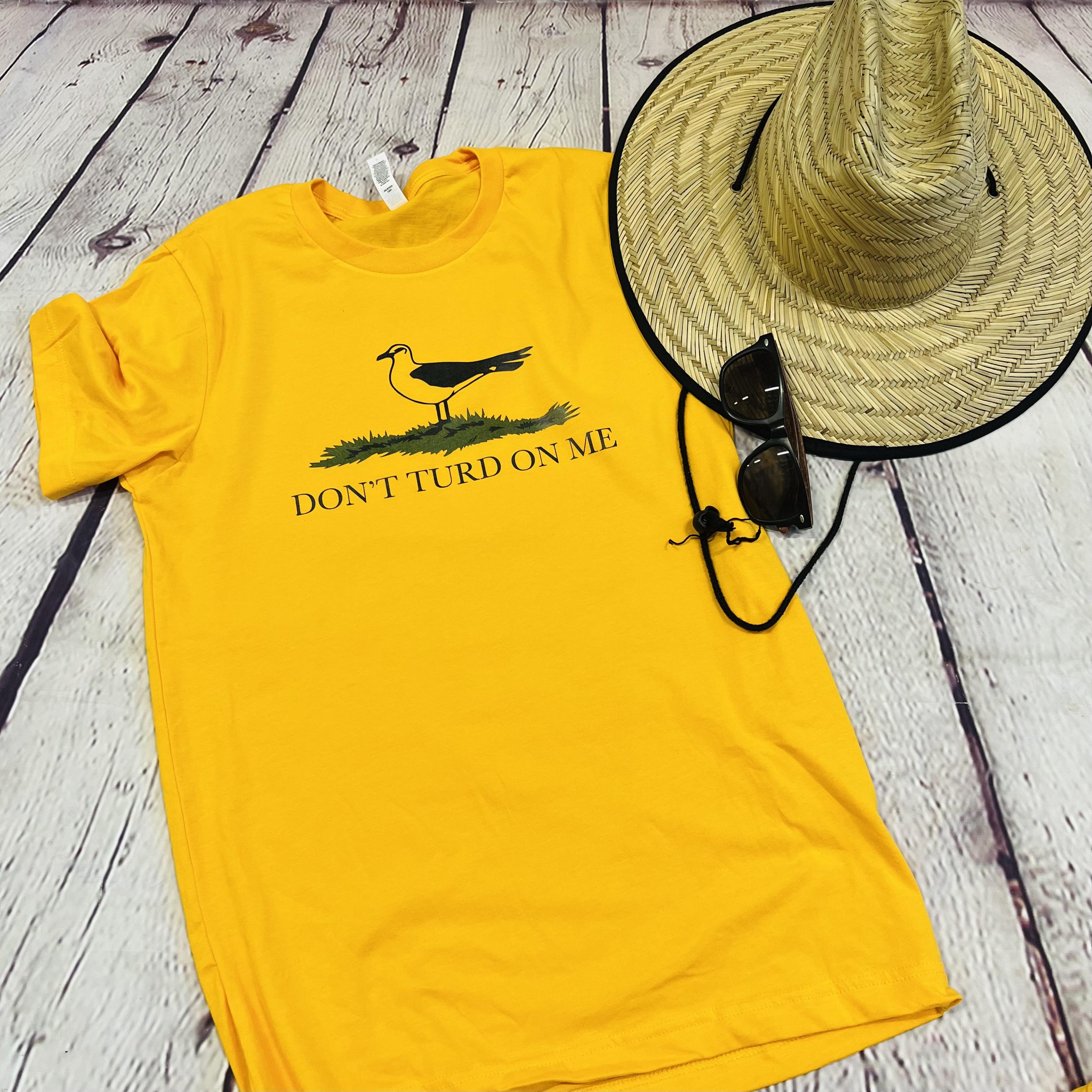 Picture of a gold yellow shirt with a seagull standing in the grass. Text on the shirt says "Don't Turd On Me."