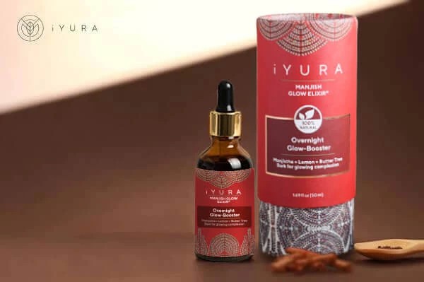 a beautiful photo of Manjish Glow Elixir - iYURA's bestselling night oil - accompanied by its beautifully illustrated, protective cylinder packaging