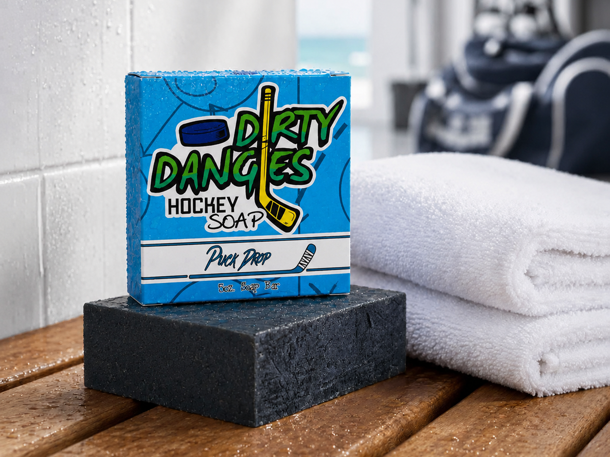Puck Drop hockey soap bar and box on a wet locker room bench beside folded white towels with hockey gear in the background.