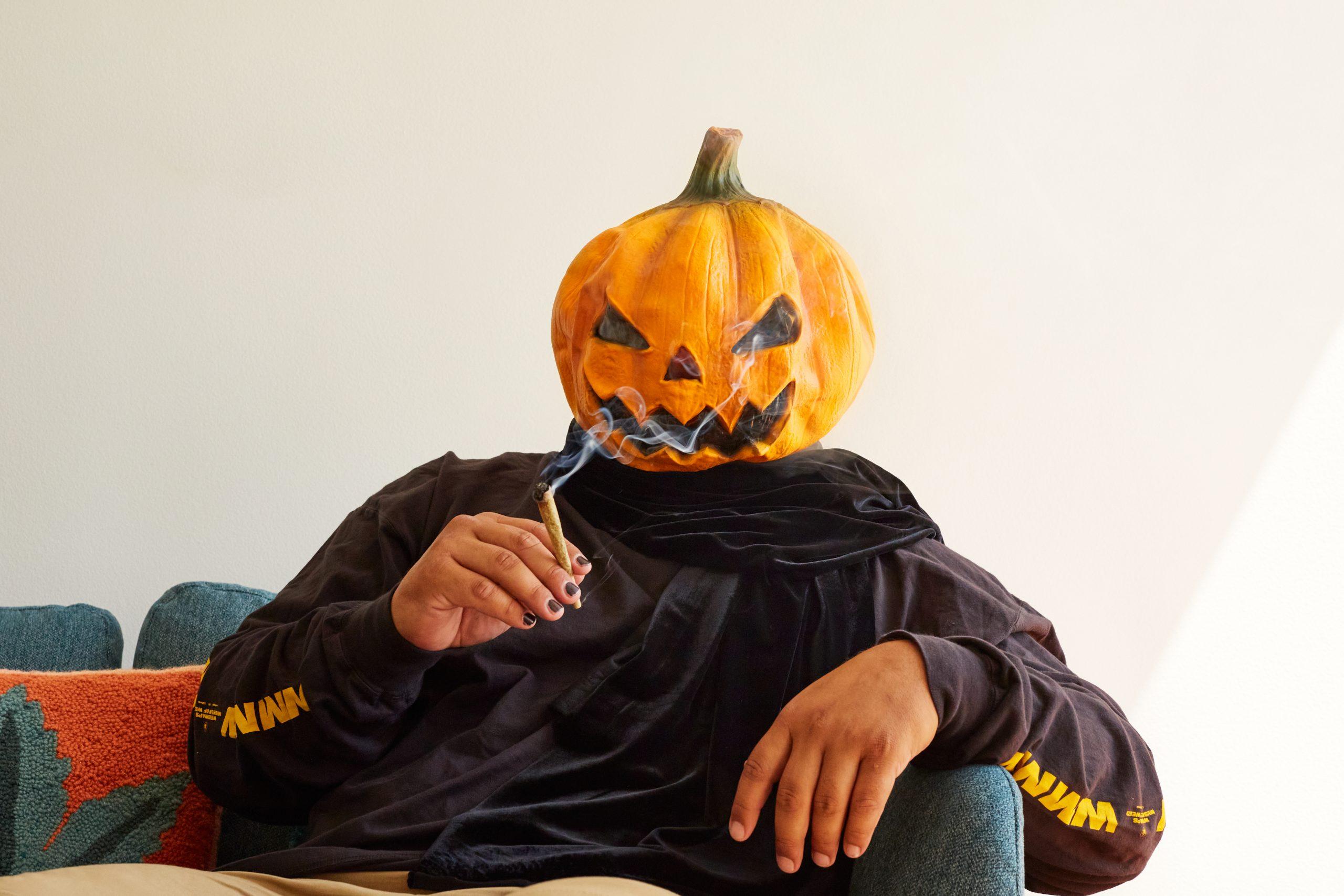 a man dressed as a jack-o'-lantern with a mask covering his face. He is reclining on a couch, relaxing, and holding a joint.