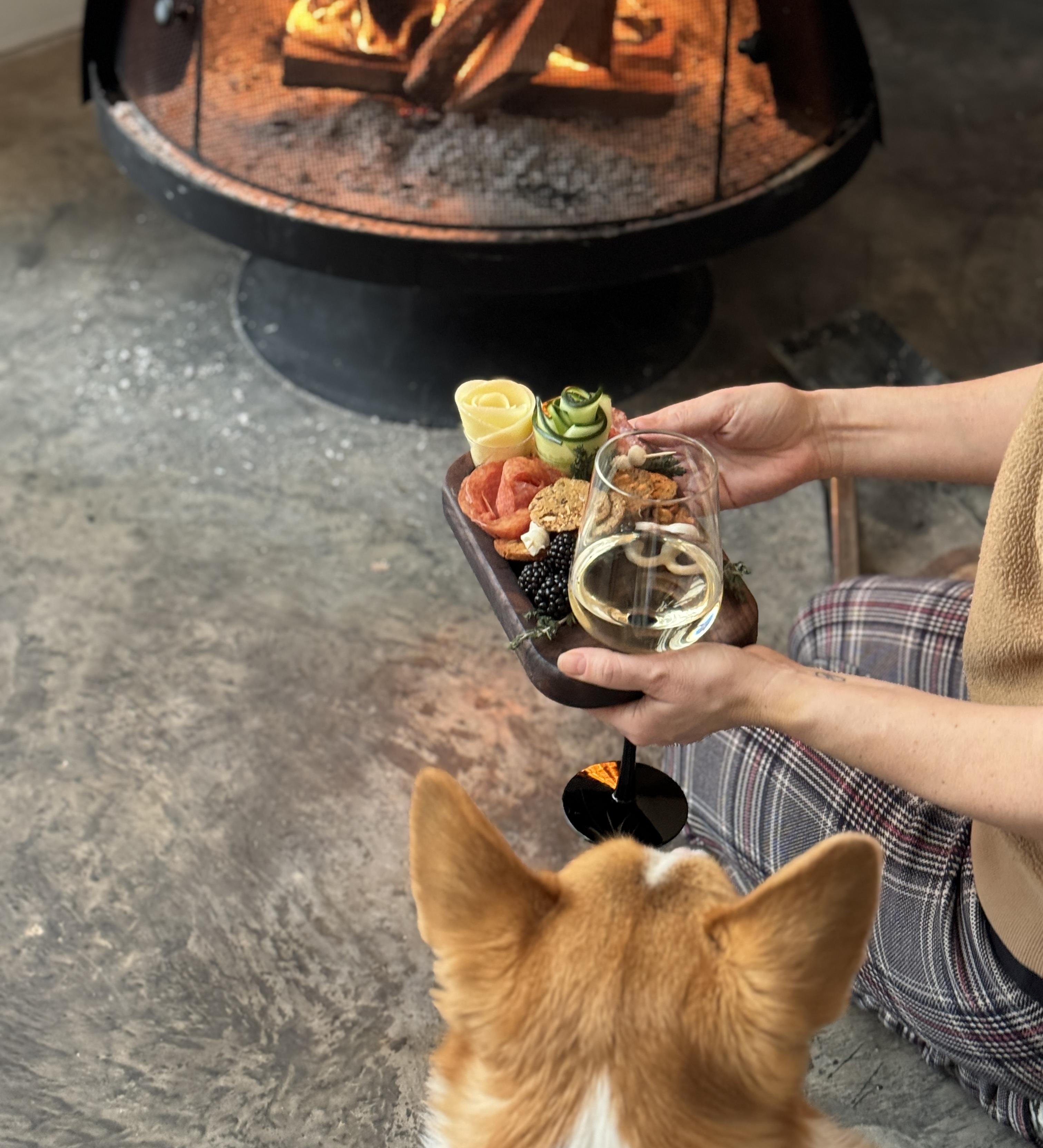 Woman holding a small personalized appetizer plate with a glass of wine and food on it in front of a fire place with a dog next to her.