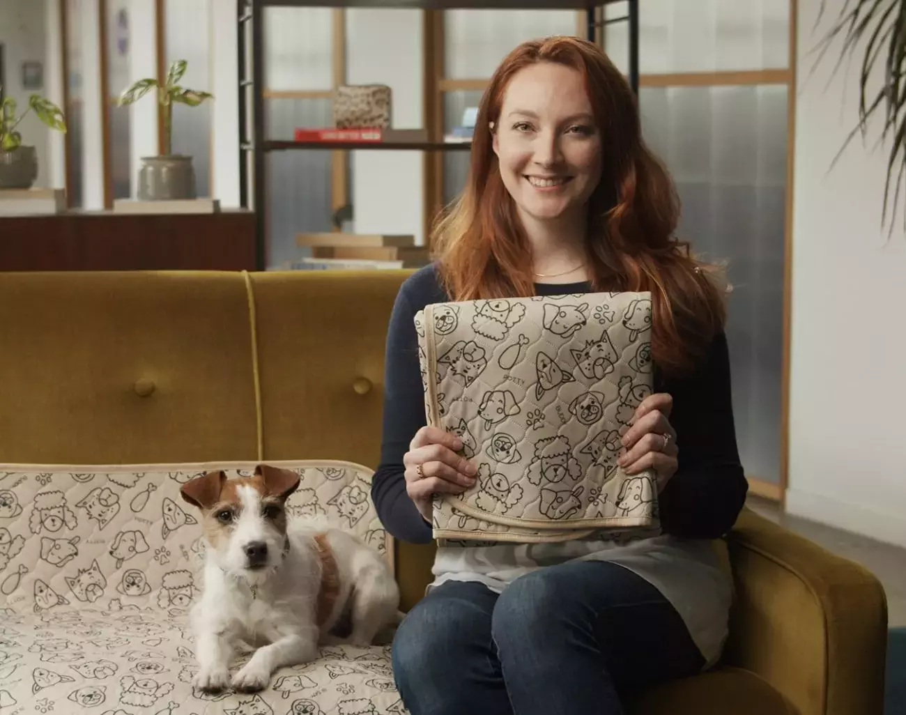 A Jack Russel dog sitting on a Potty Buddy reusable potty pad on the couch and it's owner holding up a folded pad