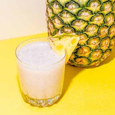 A small glass of white smoothie with a pineapple wedge set on a yellow background. Next to it is a whole pineapple fruit.