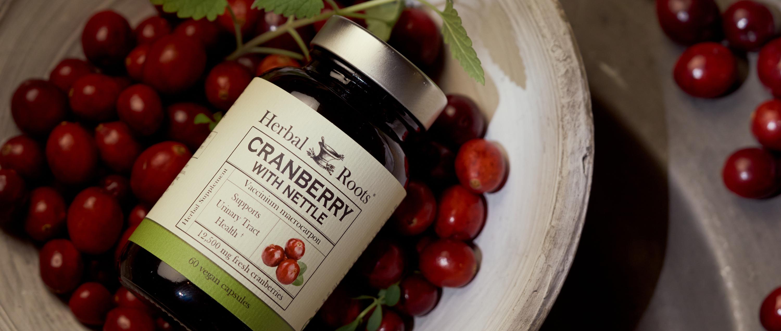 Close up aerial view of a bottle of Herbal Roots Cranberry with Nettle supplement laying face up in a bowl of fresh cranberries.