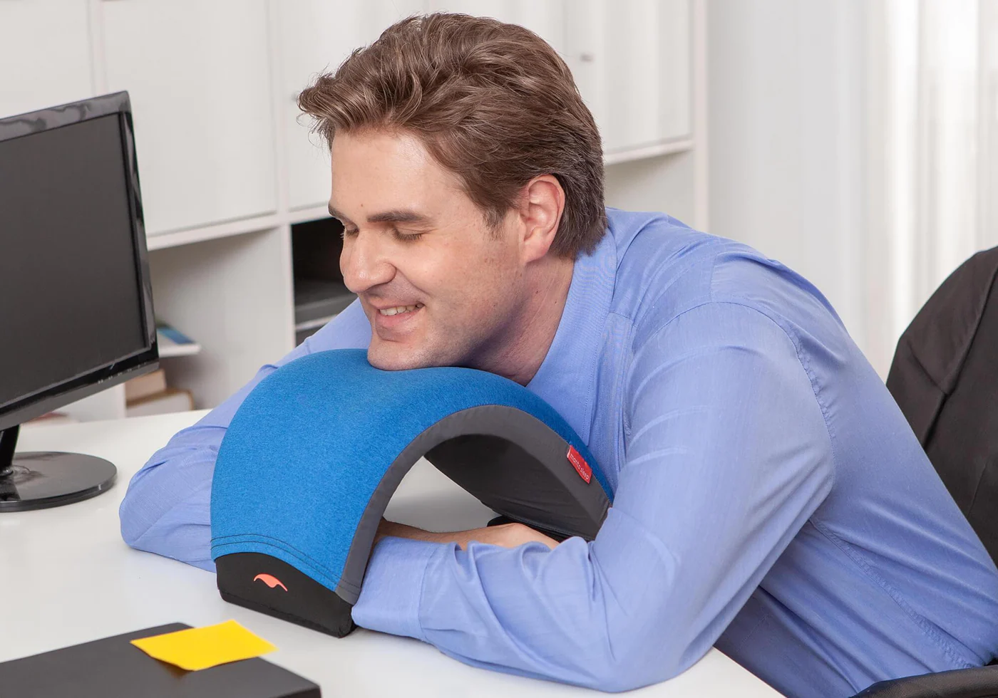 A man napping on his work desk, using a nap pillow with an arc design.