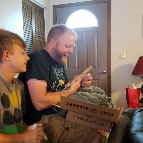 A man opening up a green dutch oven kits fart blanket gift box with his son and laughing
