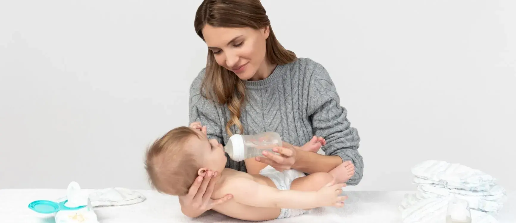 A woman in a gray sweater feeds a baby with a bottle. The baby lies on a white surface with diapers nearby. The scene conveys warmth and care.