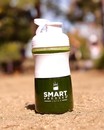 A Smart Pressed juice shaker bottle filled with green smoothie set on a sandy ground.