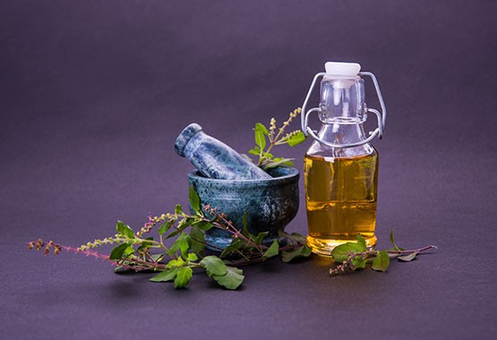 A photo of Holy Basil, oil in a glass bottle and pestle and mortar on a purple background