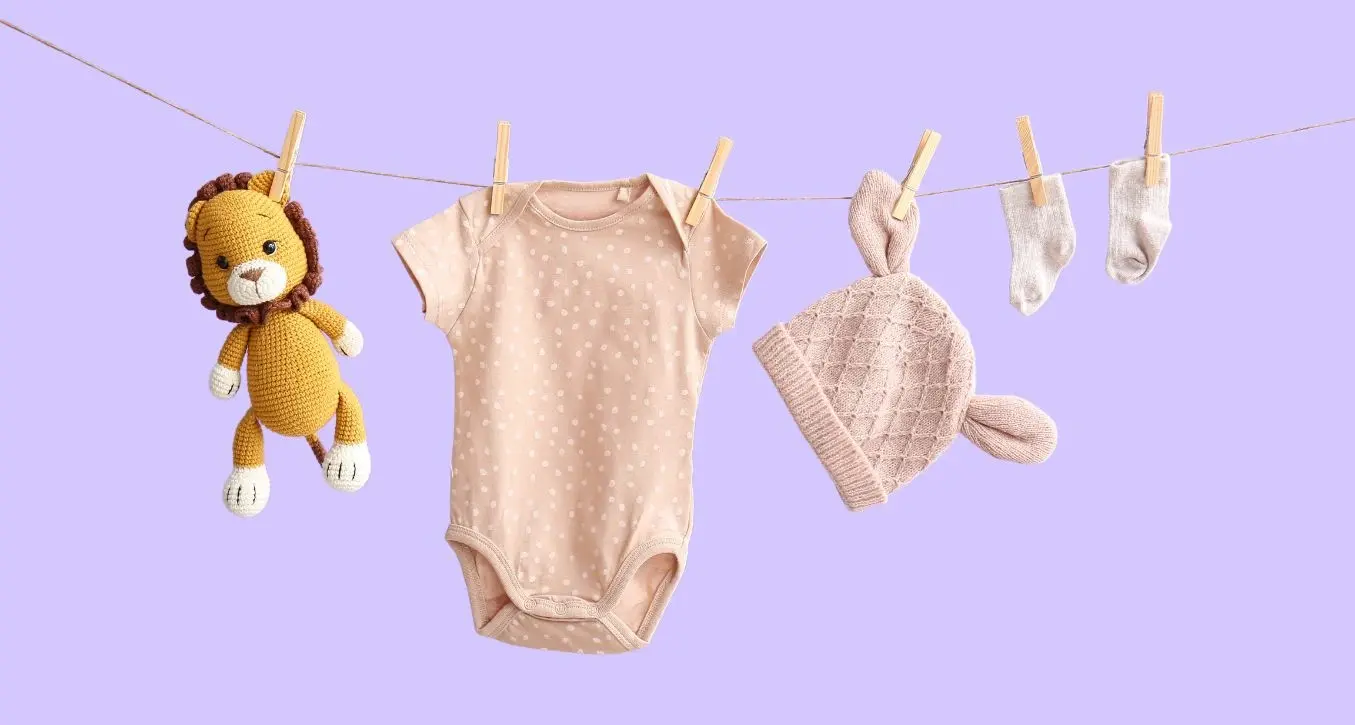 A soft pink onesie, knit hat with ears, tiny socks, and a crocheted lion toy hang on a clothesline against a pastel purple background.