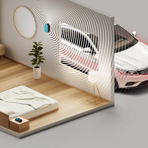 A woman sitting on a couch, holding a book. There are 2 white noise machine speakers, one on the wall and the other on a table. There is a white noise machine control pad on the table beside her.
