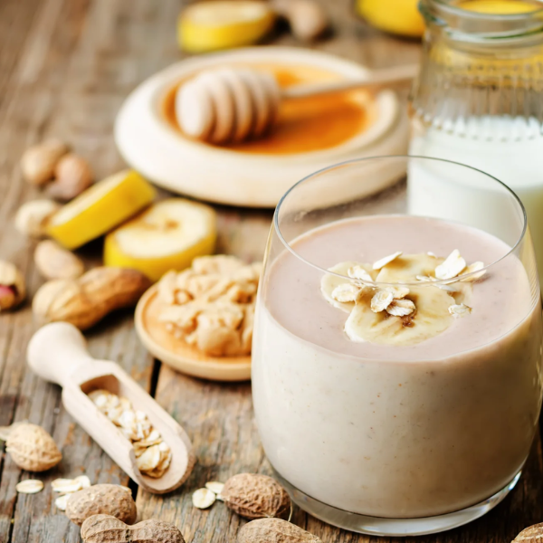 On a wooden table, there is a glass of brownish white smoothie with slices of banana and oats sprinkled on top. Behind it is a glass of milk and a plate of honey with a honey wand sitting on top. Scattered all around are slices of bananas and nuts with shells and a scooper that is half full of oats.