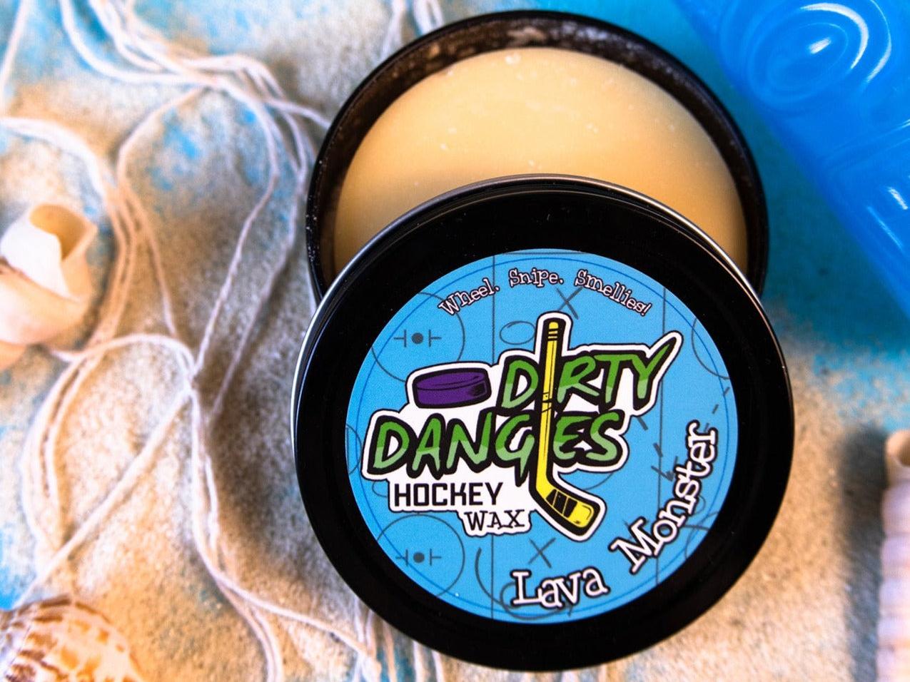 A tin of Dirty Dangles Hockey Stick Wax Lava Monster Scent sits open on a blue background surrounded by sand, seashells and a tiki mug