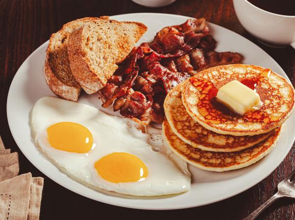 Breakfast plate with two sunny-side-up eggs, crispy bacon, toast, and a stack of pancakes topped with butter and syrup.