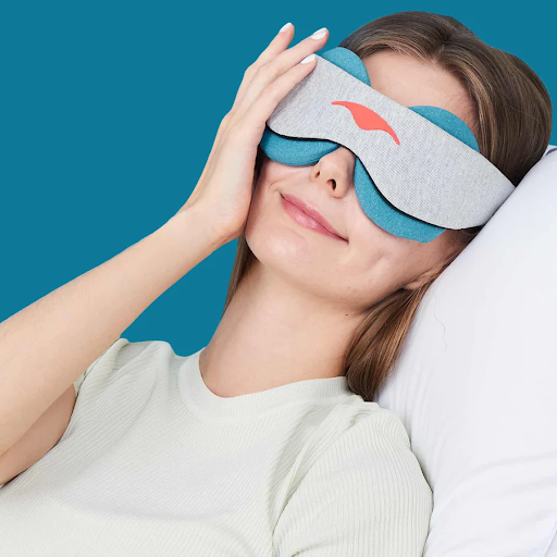 A smiling girl wearing a cooling eye mask while lying down with her hand touching her face.