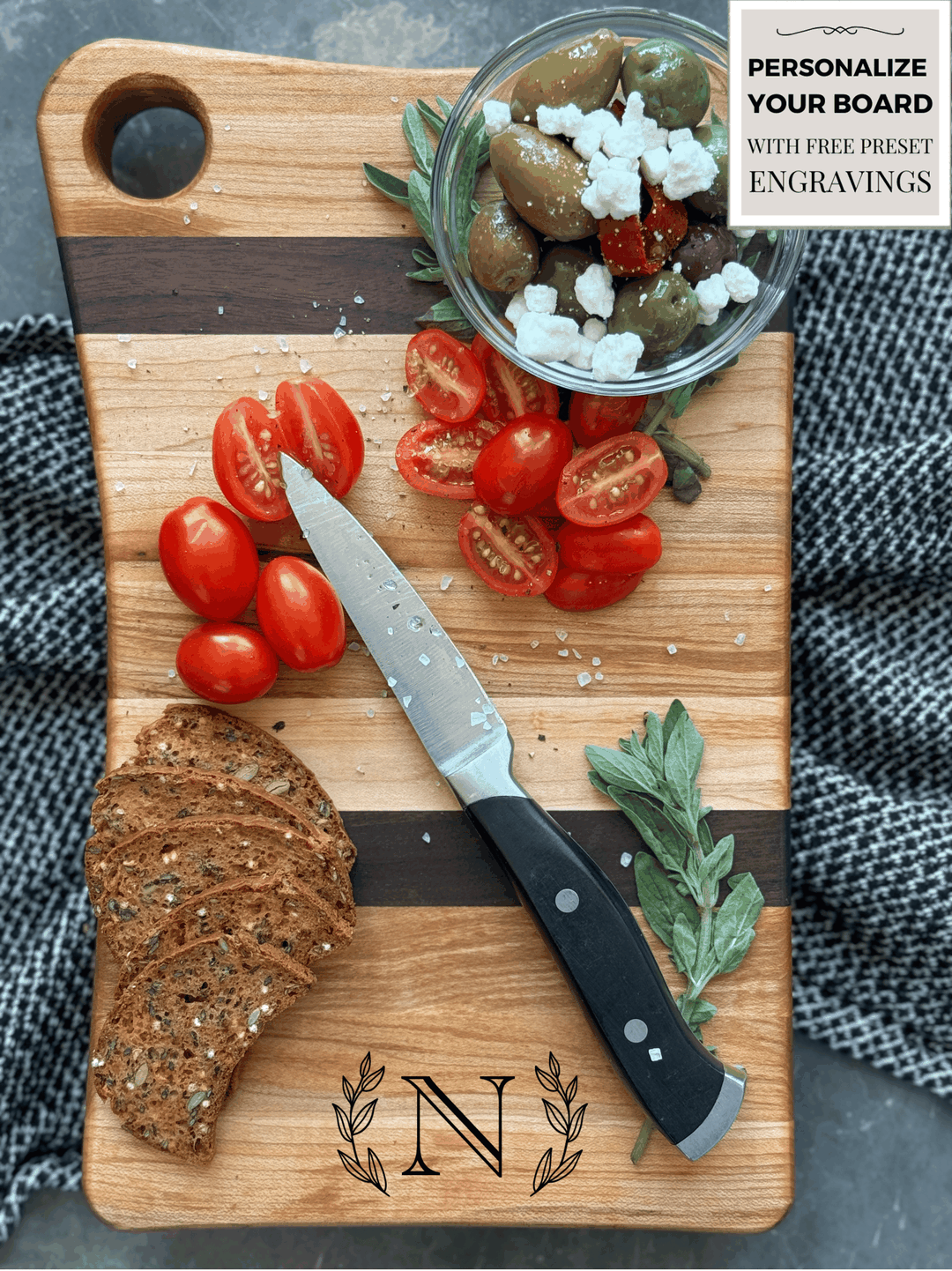 A handcrafted cutting board made of edge grain maple and walnut with a striped pattern, featuring a small round handle cutout. Topped with decorative olives, tomatoes, crackers and herbs, placed on a textured cloth background.