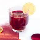 A small glass filled with red juice with a lemon wedge, set on a half red and half white surface.