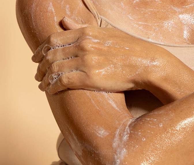 The image shows a close-up view of a woman's hands covered in a foamy, lathered body cleanser. The hands appear to be gently massaging or scrubbing the skin, indicating the use of this skincare product.