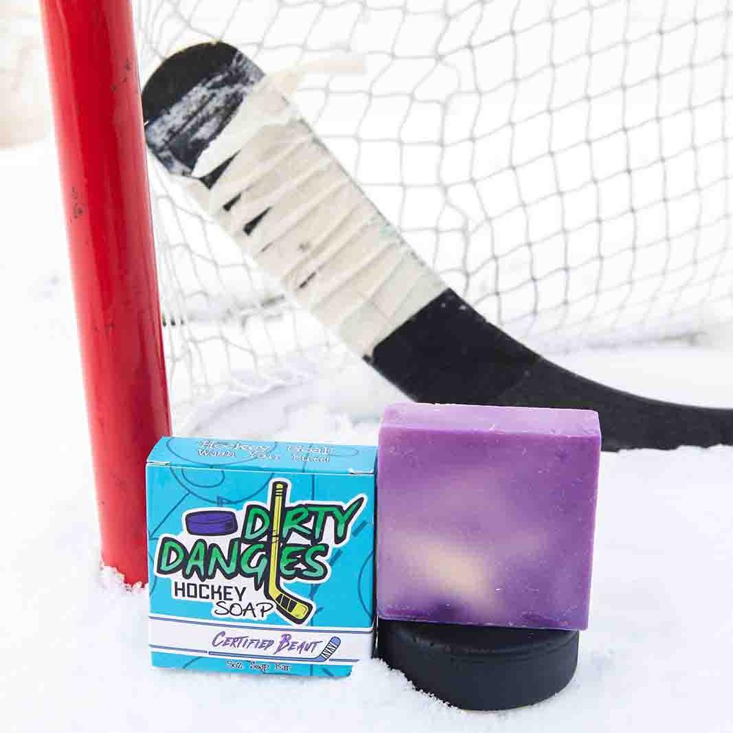 A black soap bar sits in the snow with a hockey goal, hockey puck and a hockey stick. Puck Drop