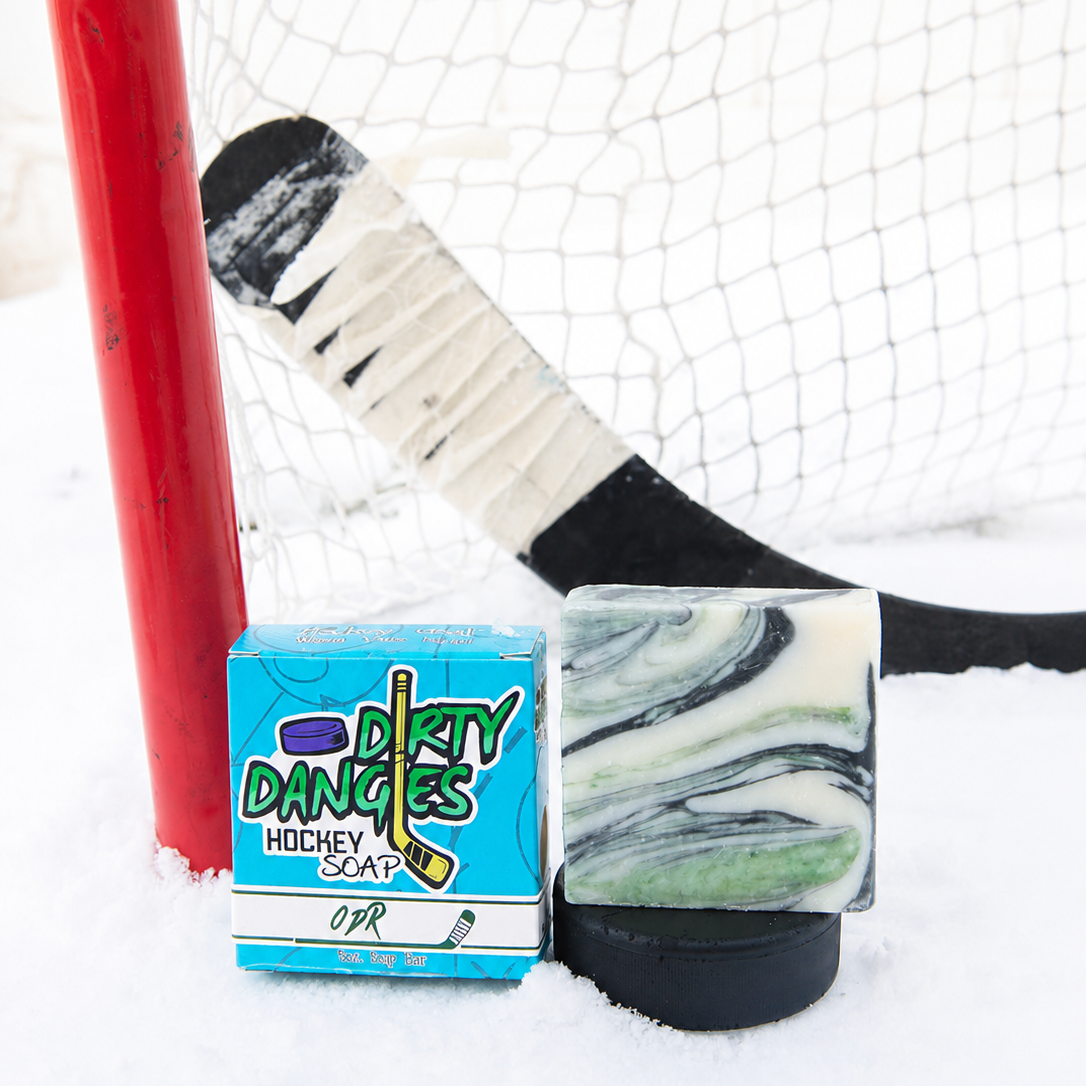 A handmade soap bar with a green and white marble pattern sits on a black hockey puck next to its blue and white 'Dirty Dangles Hockey Soap' box, set in the snow in front of a hockey goal and stick.