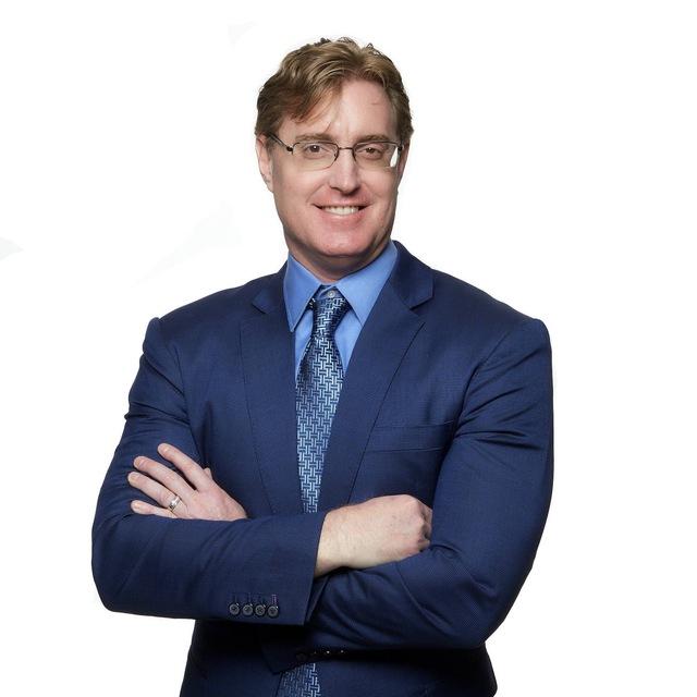 Smiling man in a blue suit and patterned tie, arms crossed