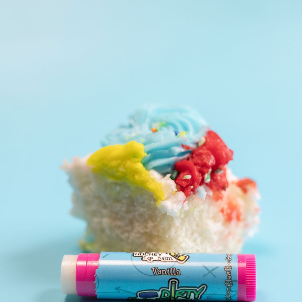 A pink stick of dirty dangles natural lip balm. Vanilla scent on a blue background with a piece of vanilla cake.