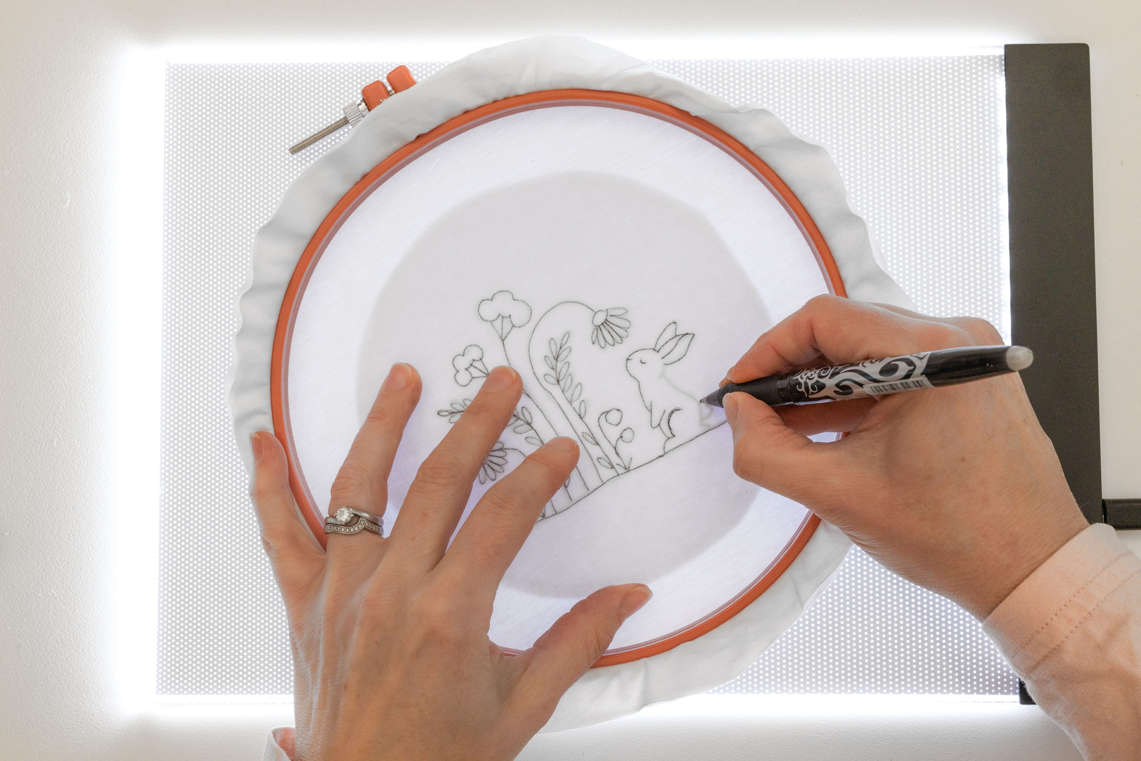 A hand draws a bunny on the fabric in a hoop using the lightbox as a backdrop.
