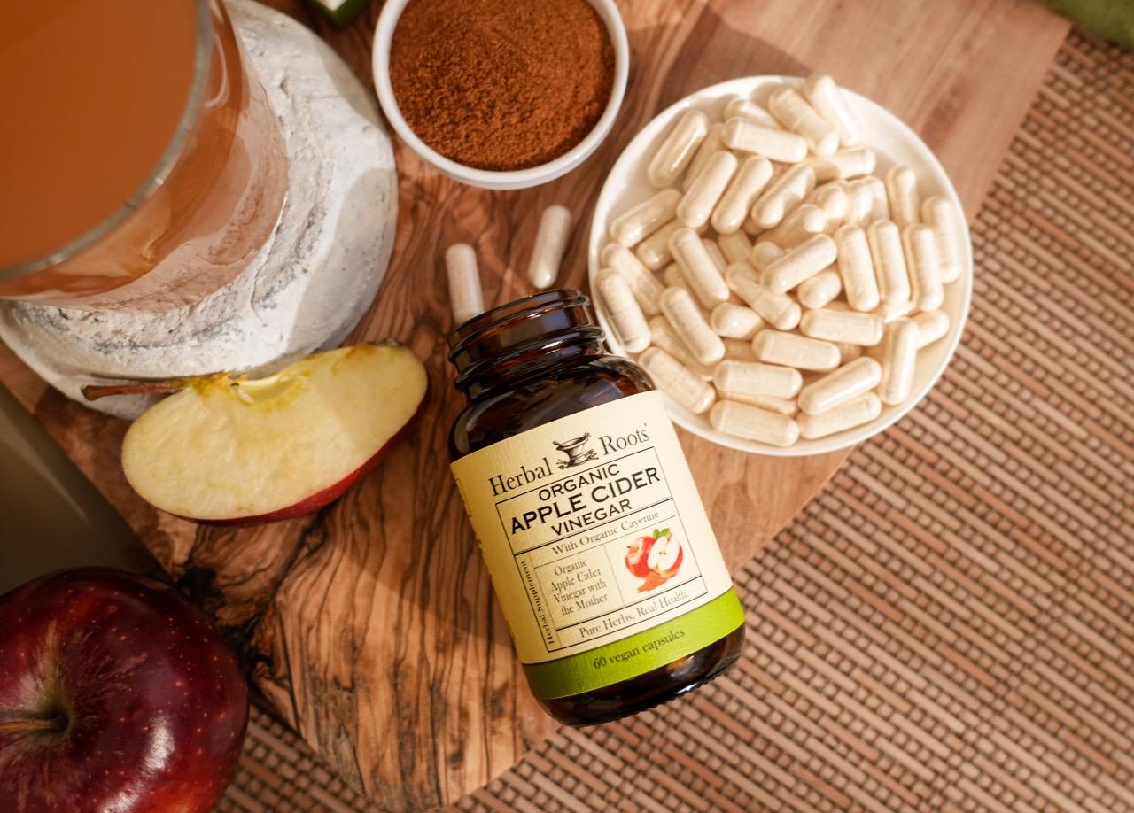 Aerial view of Herbal Roots Apple Cider Vinegar product with capsules spilling out next to a small bowl filled with capsules, a small bowl of cayenne powder, a slice of apple and a glass of ACV on a wood cutting board.