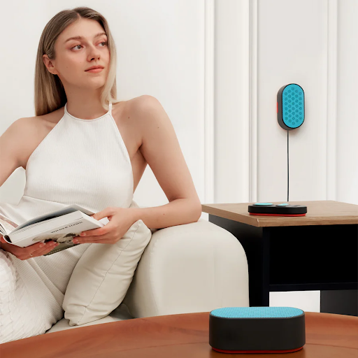 A woman sitting on a couch, holding a book. There are 2 white noise machine speakers, one on the wall and the other on a table. There is a white noise machine control pad on the table beside her.