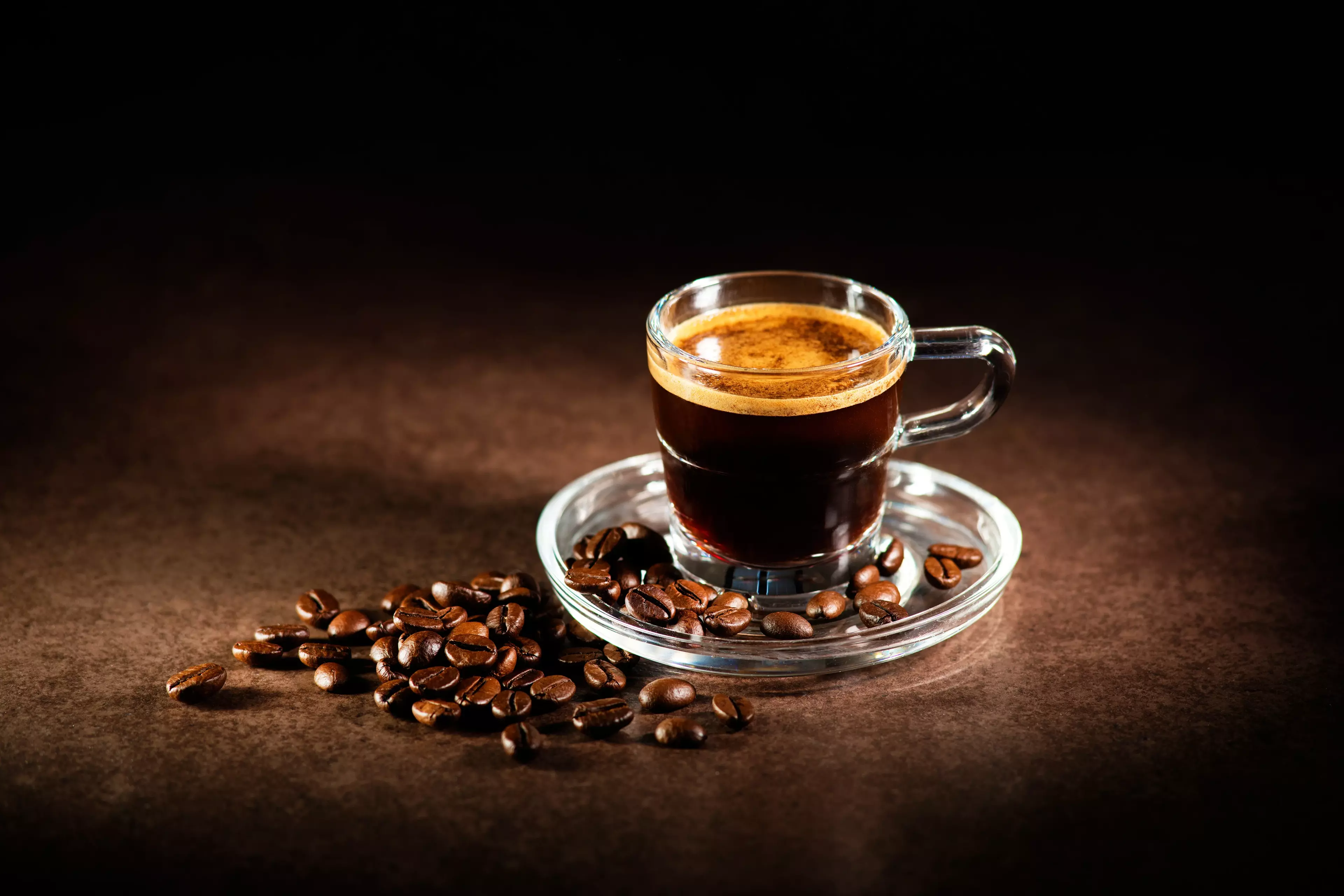Glass cup of freshly brewed espresso on a saucer surrounded by roasted coffee beans.