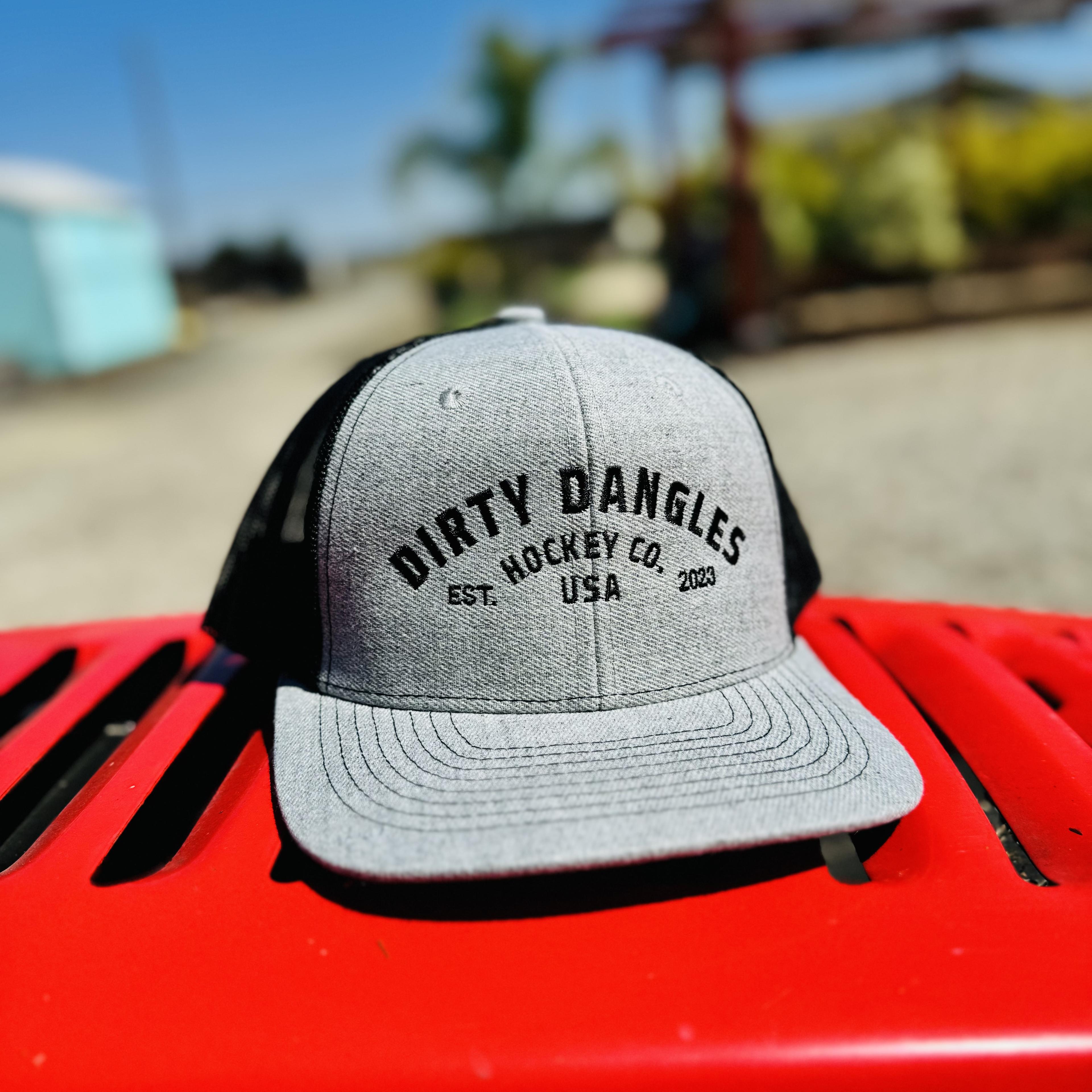 a light gray and black dirty dangles hockey co mesh snapback trucker hat outside on a red lawn mower.