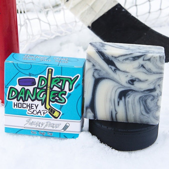 A black and white bar of dirty dangles hockey soap sits on a hockey puck in the snow with a hockey stick and a hockey goal. Sneaky Dekes Scent