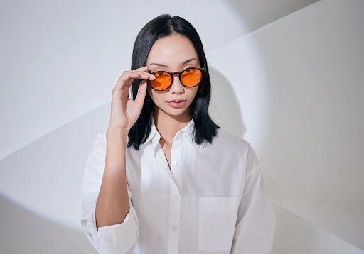 A girl in a white shirt, touching the frames of her amber-tinted blue light blocking glasses.
