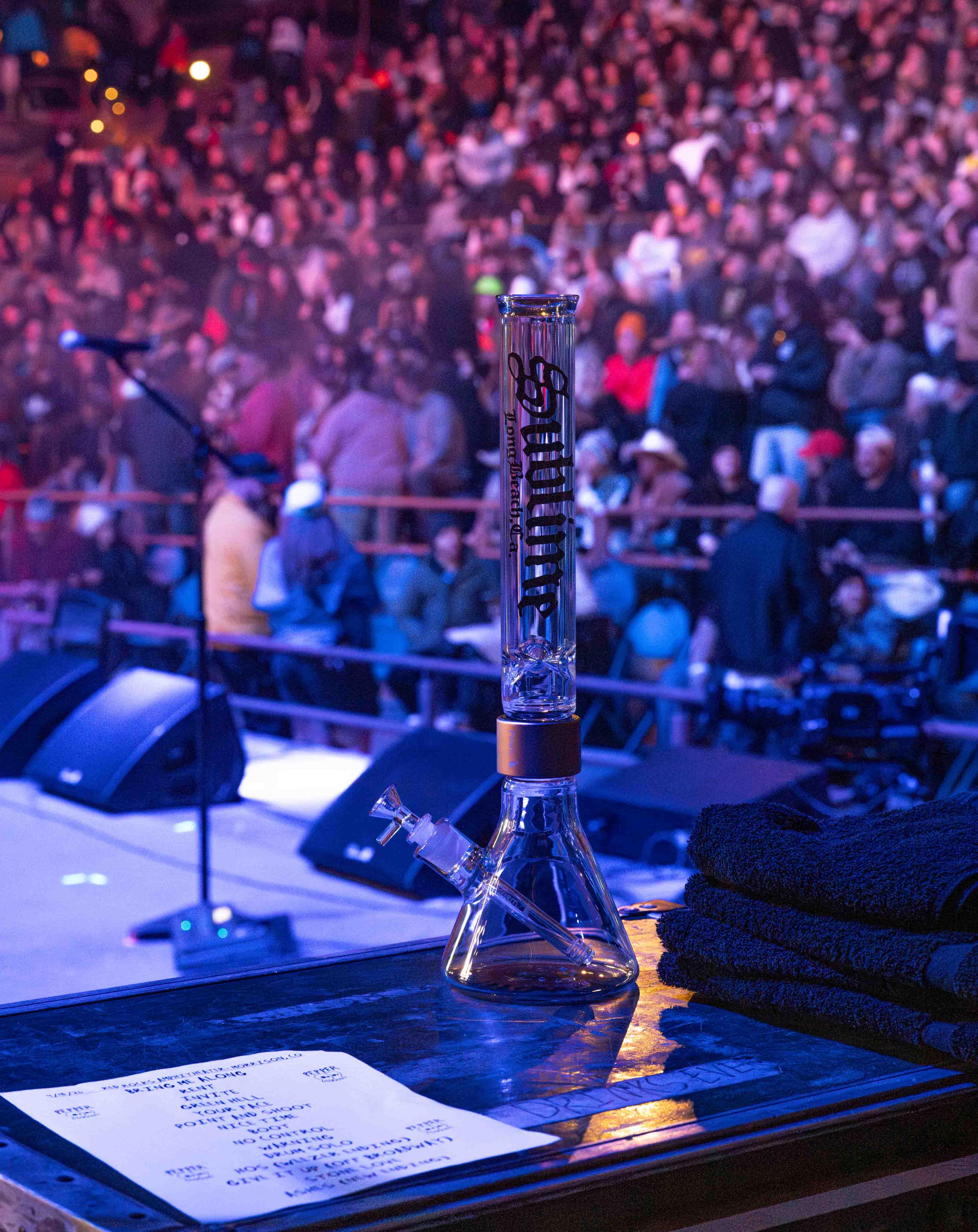 A tall clear Sublime x Prism official modular glass bong sits on the stage at Red Rocks Amphitheatre during a Sublime concert. The Red Rocks crowd is visible in the background, highlighting the official collaboration between the band and Prism during their 2026 tour.