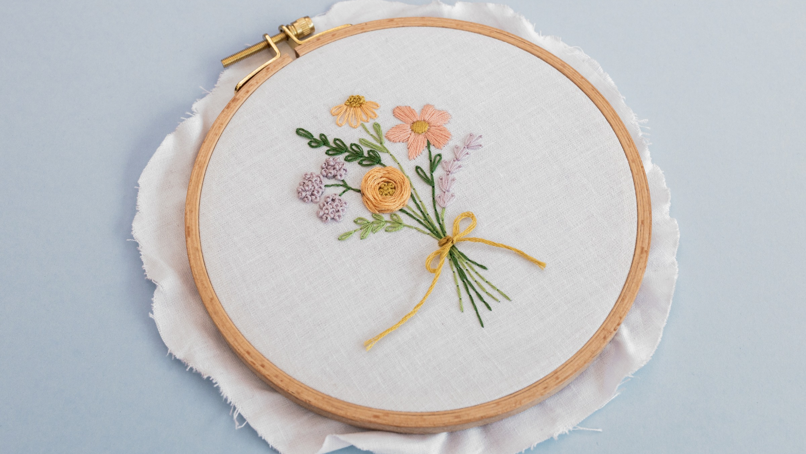 A stitched wildflower bouquet tied with a yellow bow, worked on white cotton fabric in a wooden hoop.
