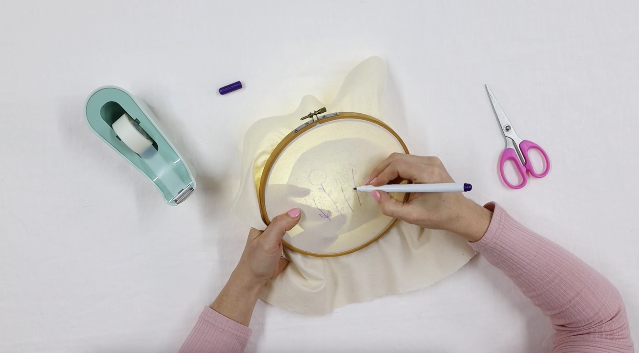 A hand transfers a pattern onto the embroidery hoop using a transfer pen.