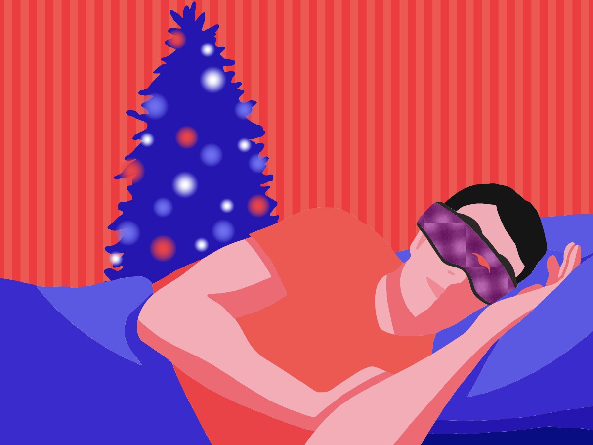 A guy wearing a sleep mask lying down with a lit Christmas tree behind him.