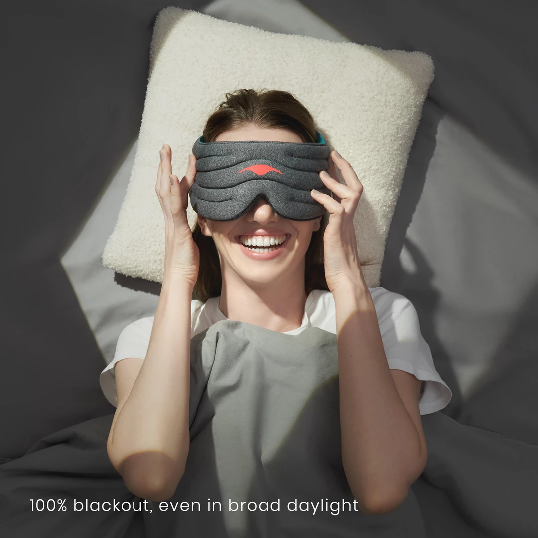 A smiling girl relaxing on her pillow touching the weighted sleep mask she is wearing.