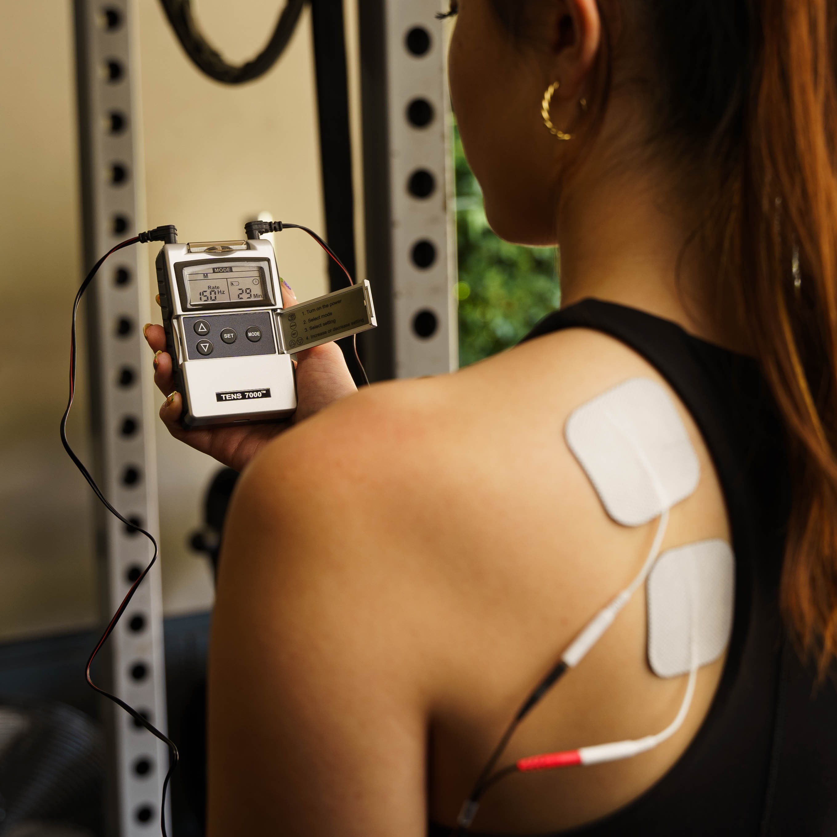 A woman holding a TENS and EMS combo unit with electrodes on her back