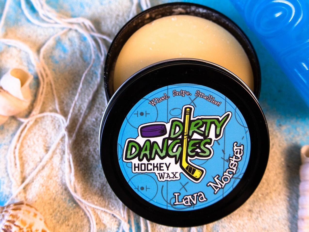 A tin of Dirty Dangles Hockey Stick Wax Lava Monster Scent sits open on a blue background surrounded by sand, seashells and a tiki mug