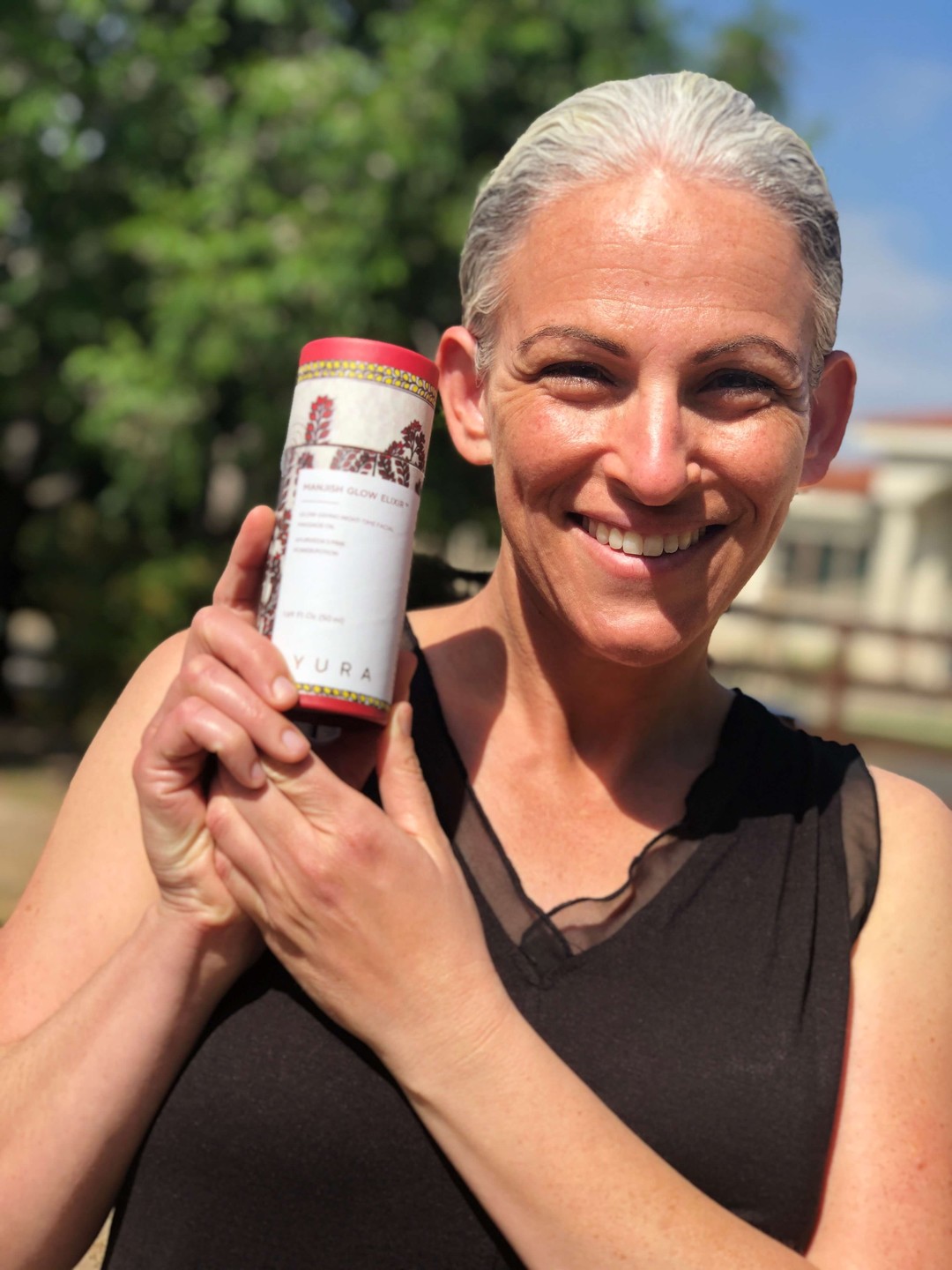 The same beautiful, peppy, mature woman holding the breathtaking, hand painted cylinder of iYURA Manjish Glow Elixir in her hand. Smiling and glowing without makeup in natural light. 