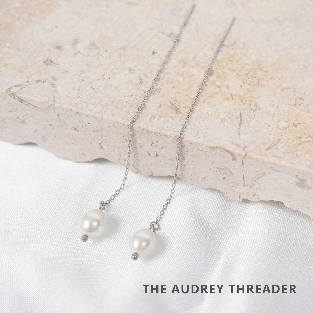 A pair of silver, hypoallergenic threader earrings with pearl drops draped over a stone surface.