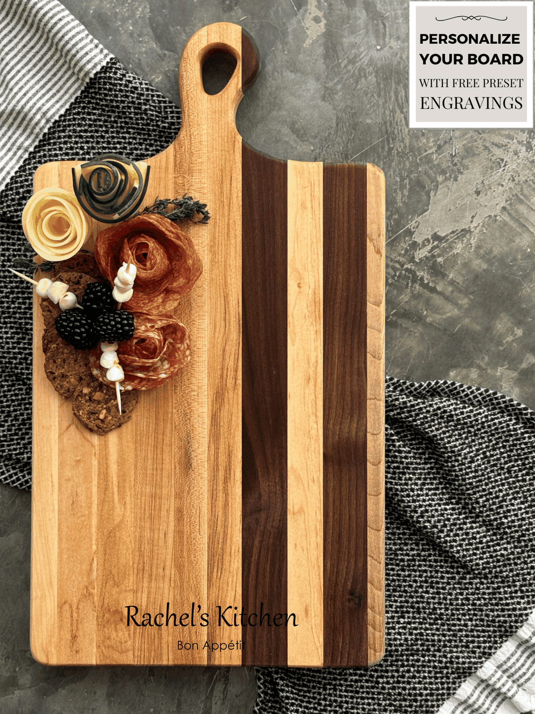 Large personalized wooden charcuterie / cutting board with Rachel Kitchen and below that, Bon Appétit engraved. Featuring elegant walnut and maple stripes, garnished with salami roses, cheese, berries, and crackers. Perfect for hosting and gifting.