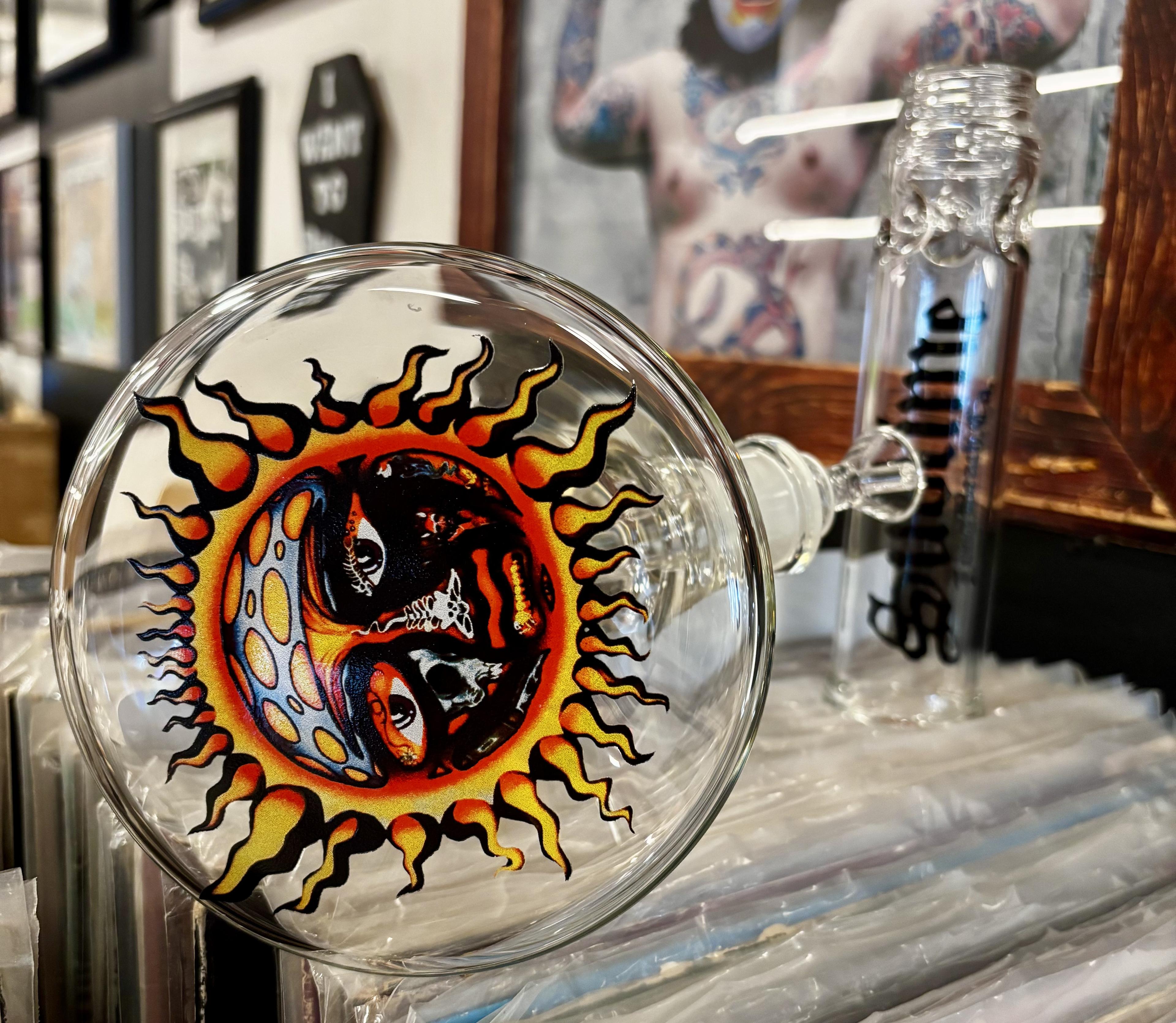 A clear Sublime x Prism official modular glass bong sits atop a stack of vinyl records in a record store. The view highlights the bottom of the beaker base, showcasing the detailed Sublime Sun logo applied to the underside of the glass.