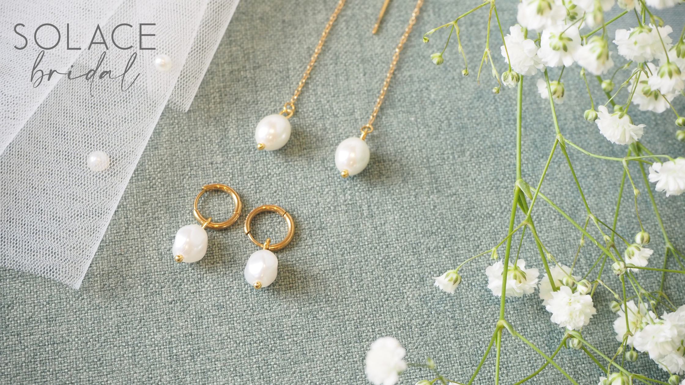 Gold hypoallergenic earrings with pearl pendants arranged on a light green fabric next to white flowers and a white veil.