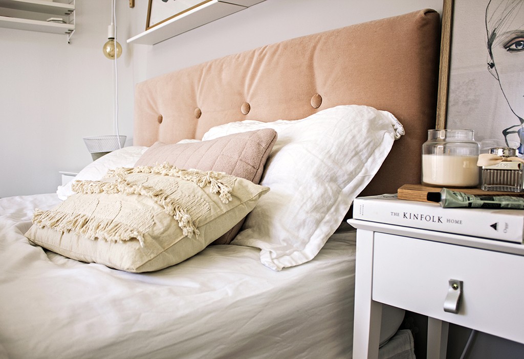 A pink upholstered headboard is on a bed surrounded by beautiful decor.