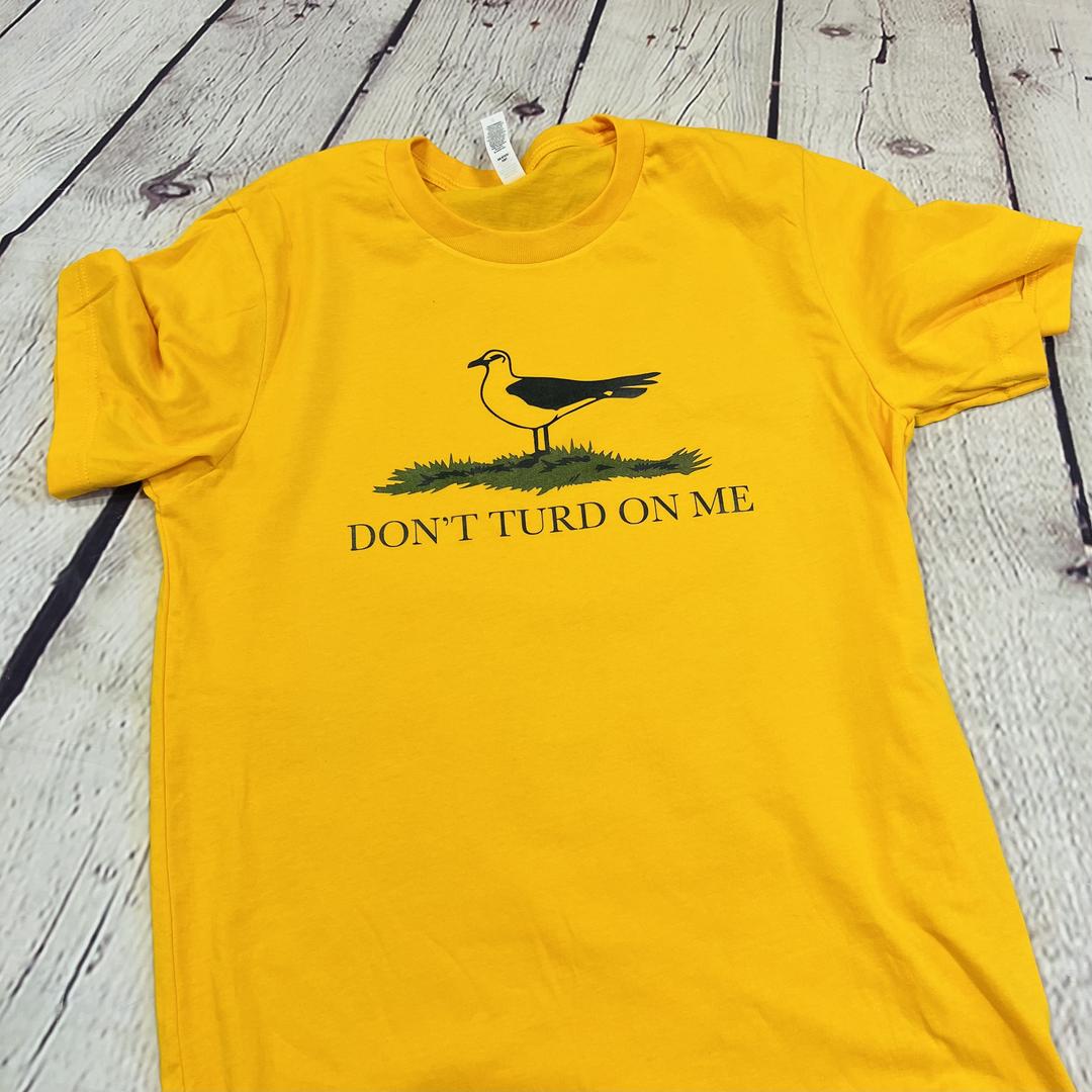 Picture of a gold and yellow t shirt with a picture of a seagull standing in the grass. Text on the shirt says "Don't Turd On Me." Similar to the Gadsden "Dont Tread On Me" Flag