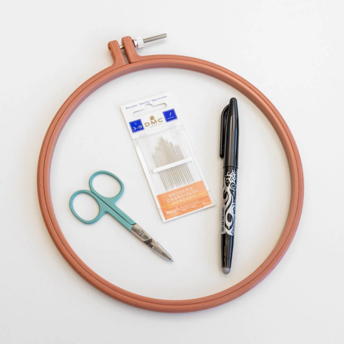 This image shows some embroidery supplies from the Embroidery Beginner's Bundle, available for purchase from the Clever Poppy Shop including Clever Poppy scissors, Mauve Rico Hoop, Pilot Frixion Pen, and DMC embroidery needles.