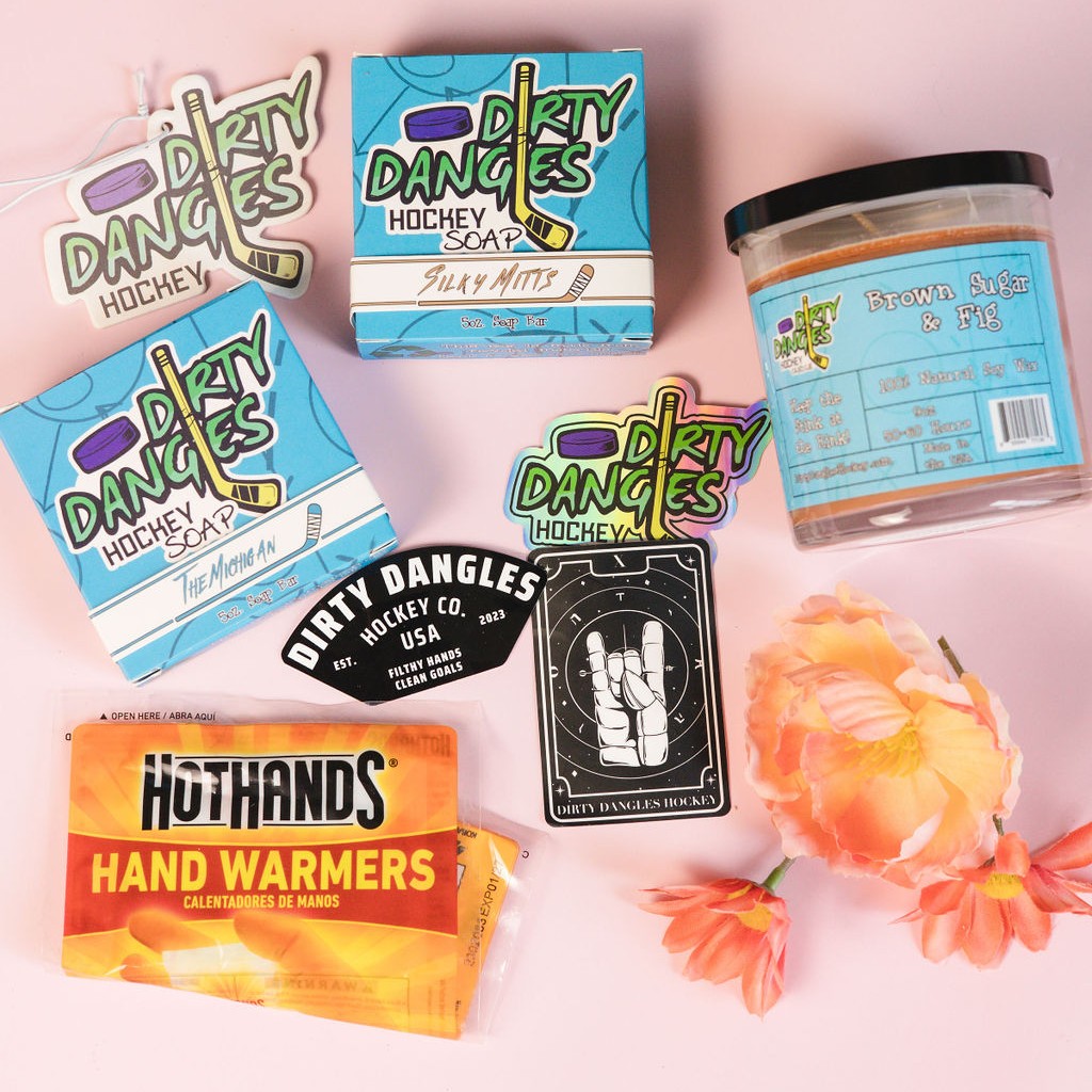 A dirty dangles hockey mom bundle on a pink background with flowers. 2 natural hocky soap bars, a scented candle, car air freshener, hand warmers and stickers