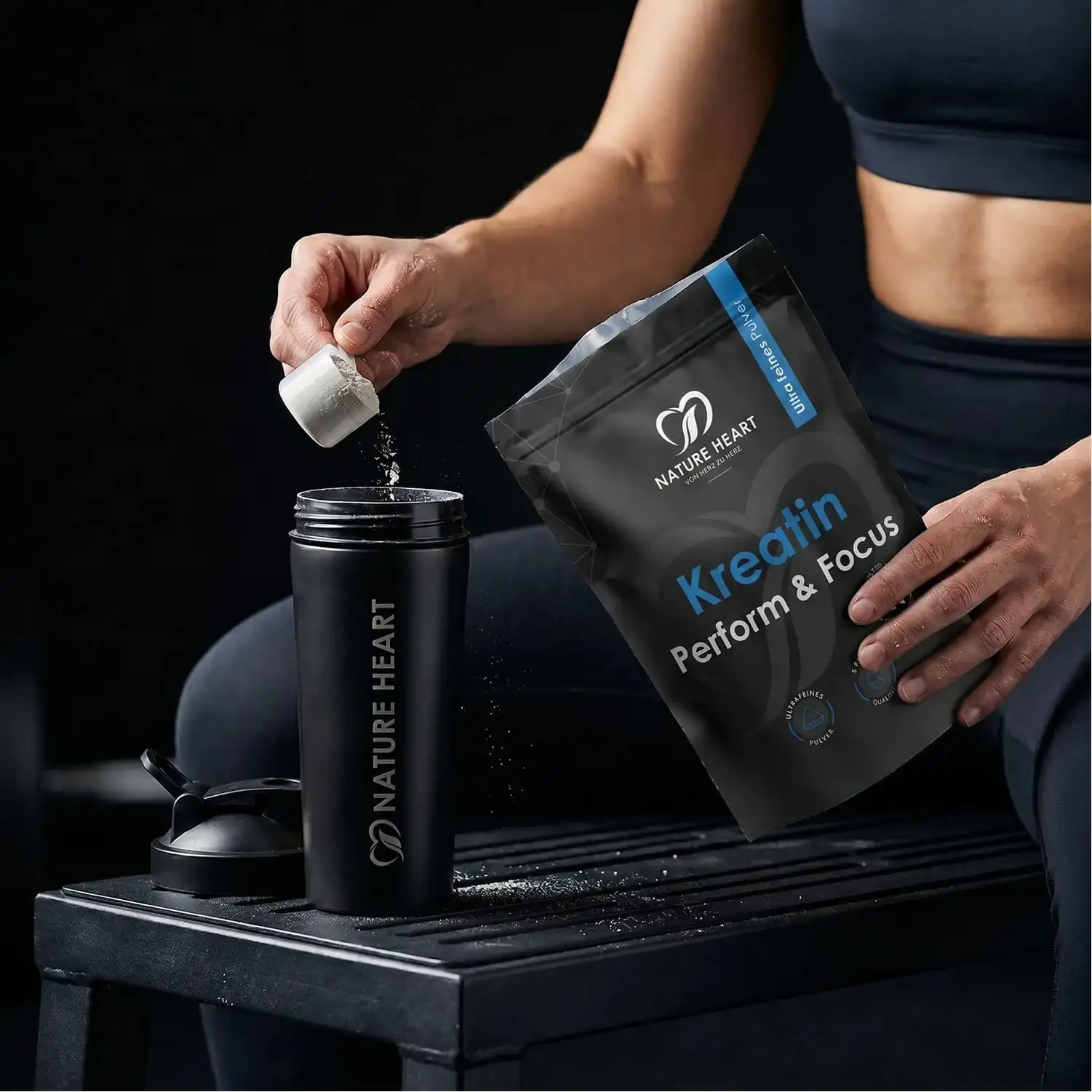 Sportlerin schüttet Nature Heart Kreatin Perform & Focus mit Messlöffel in einen Nature Heart Shaker im dunklen Gym